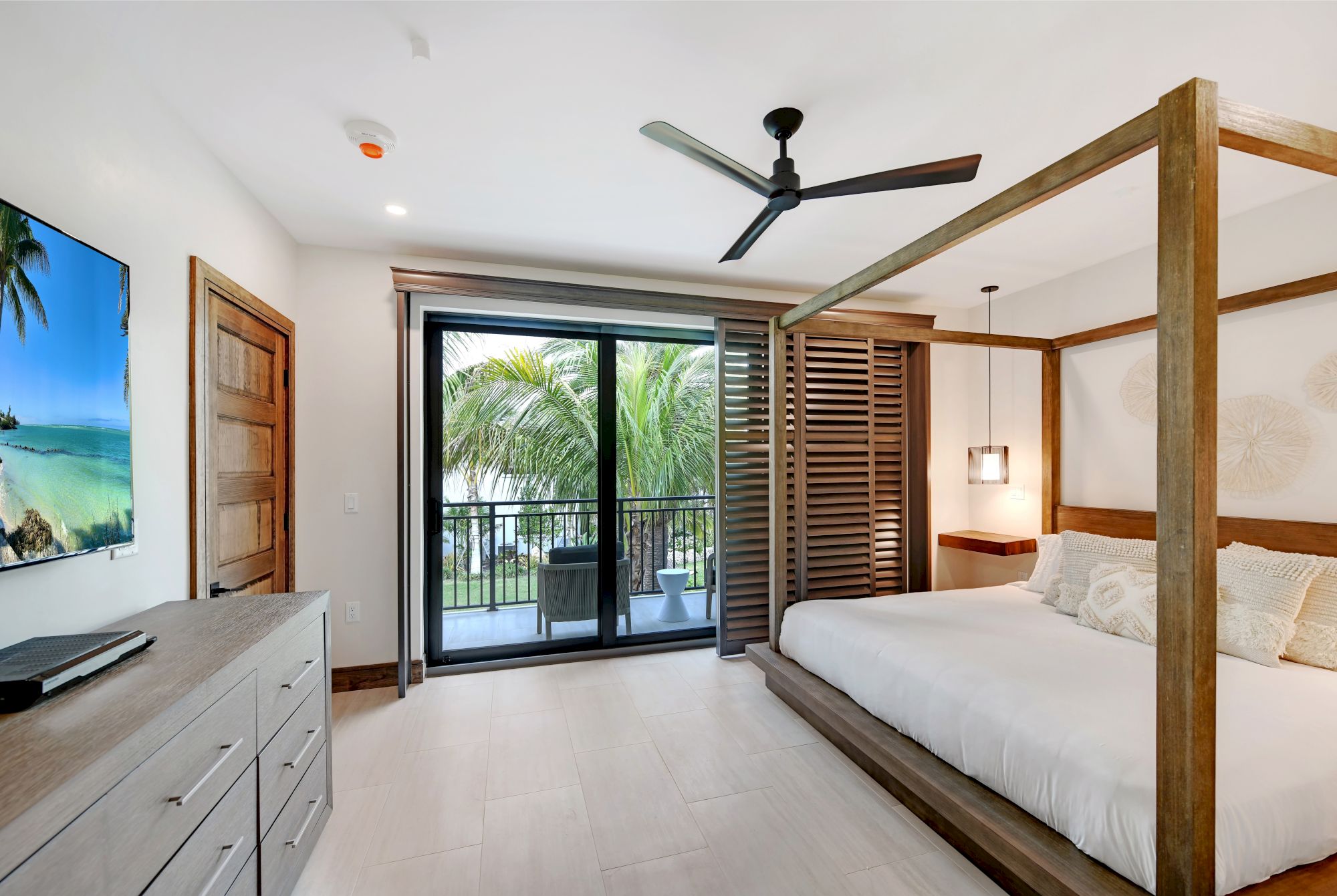 This image shows a modern bedroom with a canopy bed, dresser, TV, ceiling fan, and a sliding door leading to a balcony with a view of palm trees.