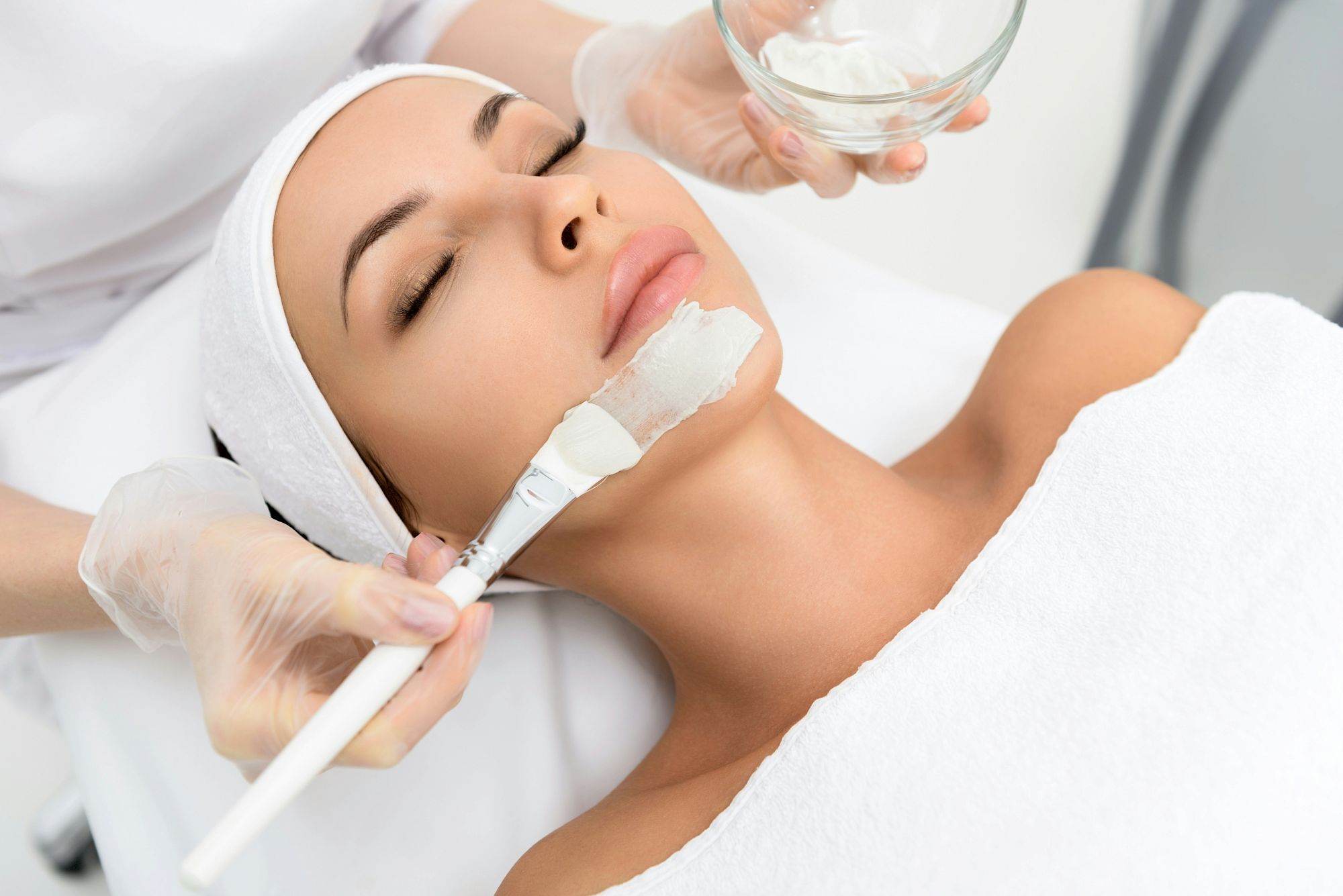 A woman is receiving a facial treatment with a mask being applied to her face using a brush by a practitioner.