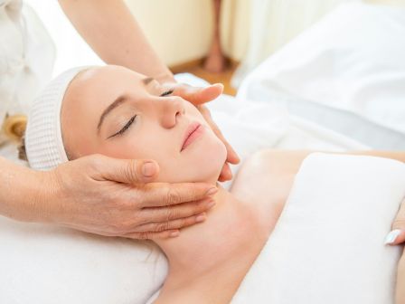 A person is receiving a facial massage while lying down with their eyes closed, covered in a white towel and headband in a serene environment.