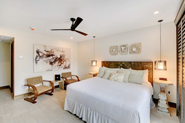 This image shows a modern bedroom with a large bed, two chairs, a ceiling fan, and wall art. The decor is minimal and elegant.