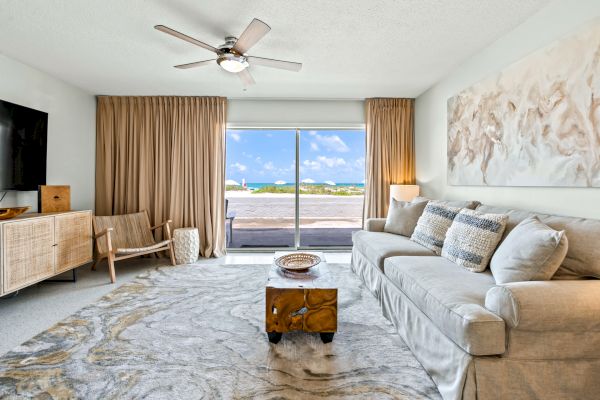 A modern living room with a large sofa, wooden coffee table, armchair, TV, and a glass door leading to a beautiful outdoor view of the ocean.