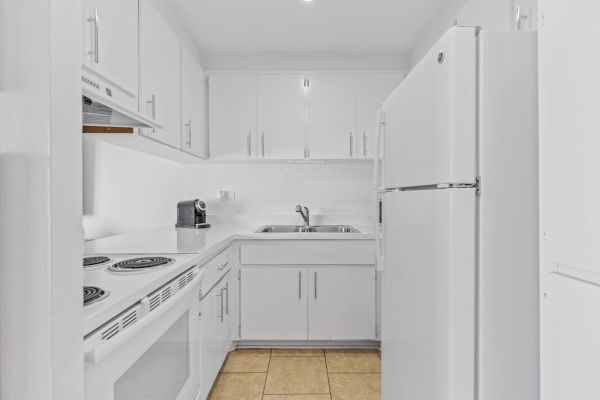 A modern white kitchen with cabinets, a sink, stove, refrigerator, and a small coffee machine on the counter. The floor has beige tiles.