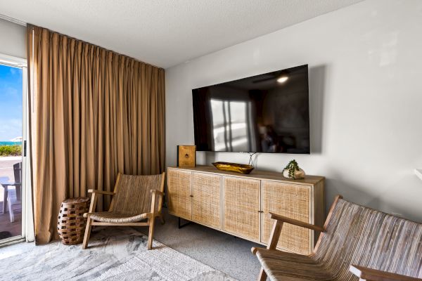 The image shows a modern living room with a wall-mounted TV, two wicker chairs, a light brown rug, and a wooden cabinet under the TV.