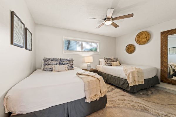 A cozy bedroom with two beds, stylish decor, a ceiling fan, wall art, and a large mirror. A window provides natural light.