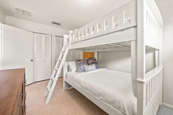 A bedroom with white bunk beds, featuring a ladder and blue-patterned pillows, beside a wooden dresser and partially open closet doors.