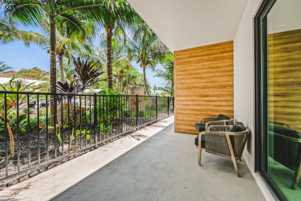 A tropical patio with two chairs, wooden wall accents, potted plants, and a view of lush greenery, enclosed by a black railing.