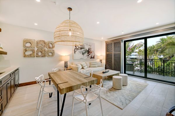 A modern open-concept living area with dining table, sofa, artwork, pendant lamp, and large sliding doors leading to a balcony with greenery outside.