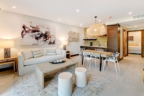 The image shows a modern living room and kitchen area with stylish furniture, a couch, dining table, and wall art, leading to a bedroom.