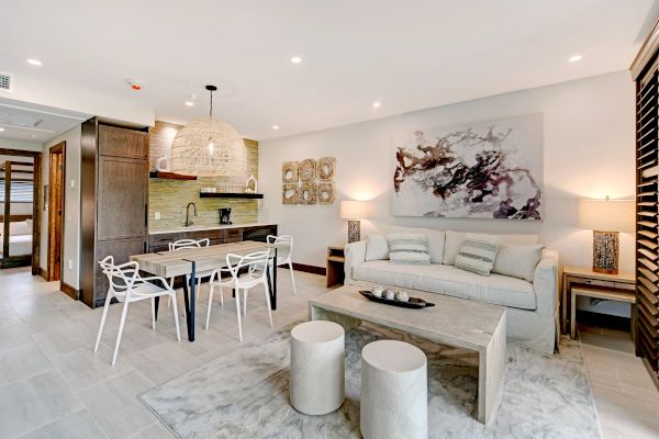 A modern living area with a sofa, dining table, and kitchenette. Decor includes abstract art, pendant light, and stools on a rug, ending the sentence.