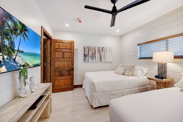 A modern bedroom with two beds, a wall-mounted TV displaying a beach scene, a ceiling fan, and stylish decor, ending the sentence.