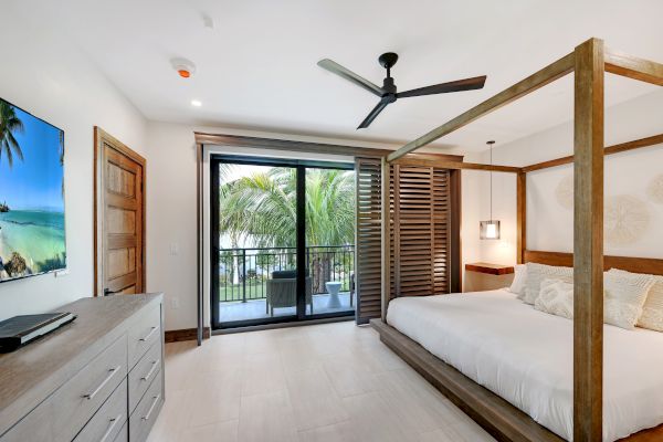 A modern bedroom with a canopy bed, ceiling fan, flat-screen TV, dresser, and sliding doors leading to a balcony with a tropical view.