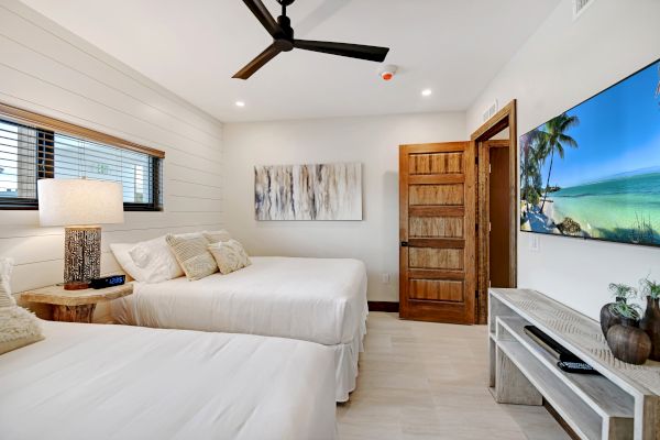 A modern bedroom with two beds, a large flat-screen TV, a ceiling fan, wooden accents, art, and ample natural light from a window.