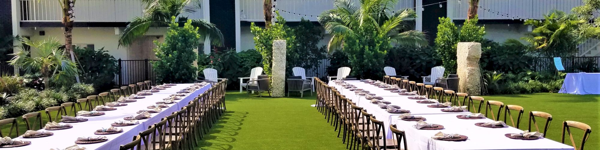 An outdoor dining setup with two rows of tables and chairs, surrounded by palm trees and greenery in front of a building.