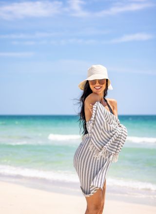 A smiling person wearing a striped dress and sun hat is posing on a beach with a blue ocean and sky in the background.