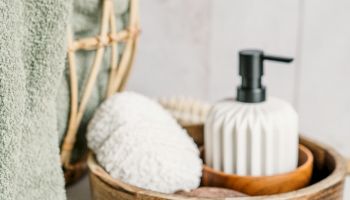 The image shows a spa-like setup with a green towel, a wicker basket, a soap dispenser, a sponge, a wooden bowl, and a lit candle on a white surface.