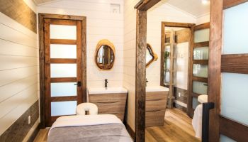 A cozy interior room with a massage table, wooden doors, mirrors, sinks, and modern lighting. The design features a mix of white shiplap and wood.