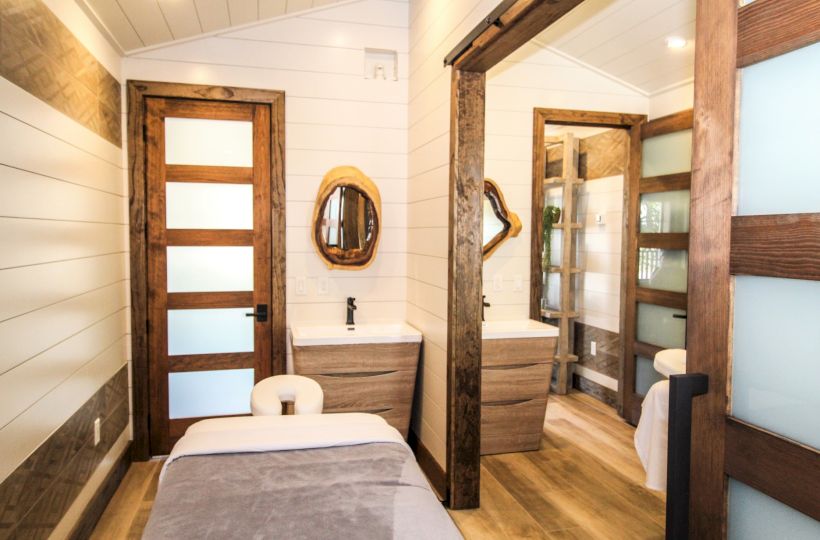 A cozy interior room with a massage table, wooden doors, mirrors, sinks, and modern lighting. The design features a mix of white shiplap and wood.