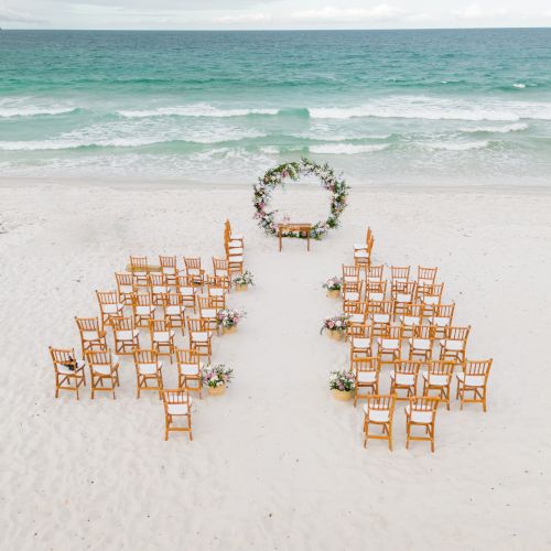 An outdoor wedding setup on a beach with wooden chairs arranged in rows facing a round floral arch, with the ocean in the background.