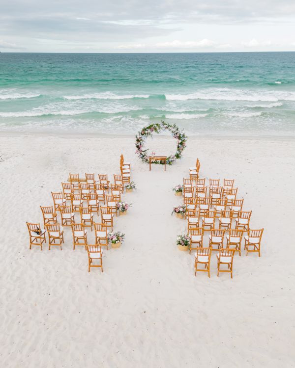 An outdoor wedding setup on a beach with wooden chairs arranged in rows facing a round floral arch, with the ocean in the background.