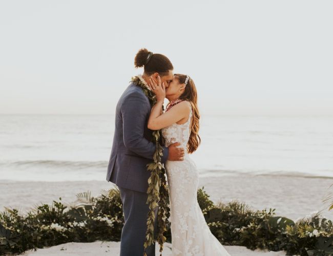 A couple shares a kiss in a beachside wedding ceremony, surrounded by greenery and ocean waves in the background.