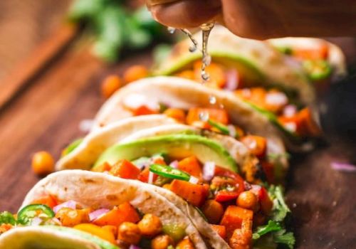 The image shows a person squeezing lime juice onto a row of colorful tacos filled with chickpeas and vegetables, displayed on a wooden board.