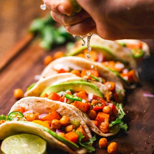 The image shows a person squeezing lime juice onto a row of colorful tacos filled with chickpeas and vegetables, displayed on a wooden board.