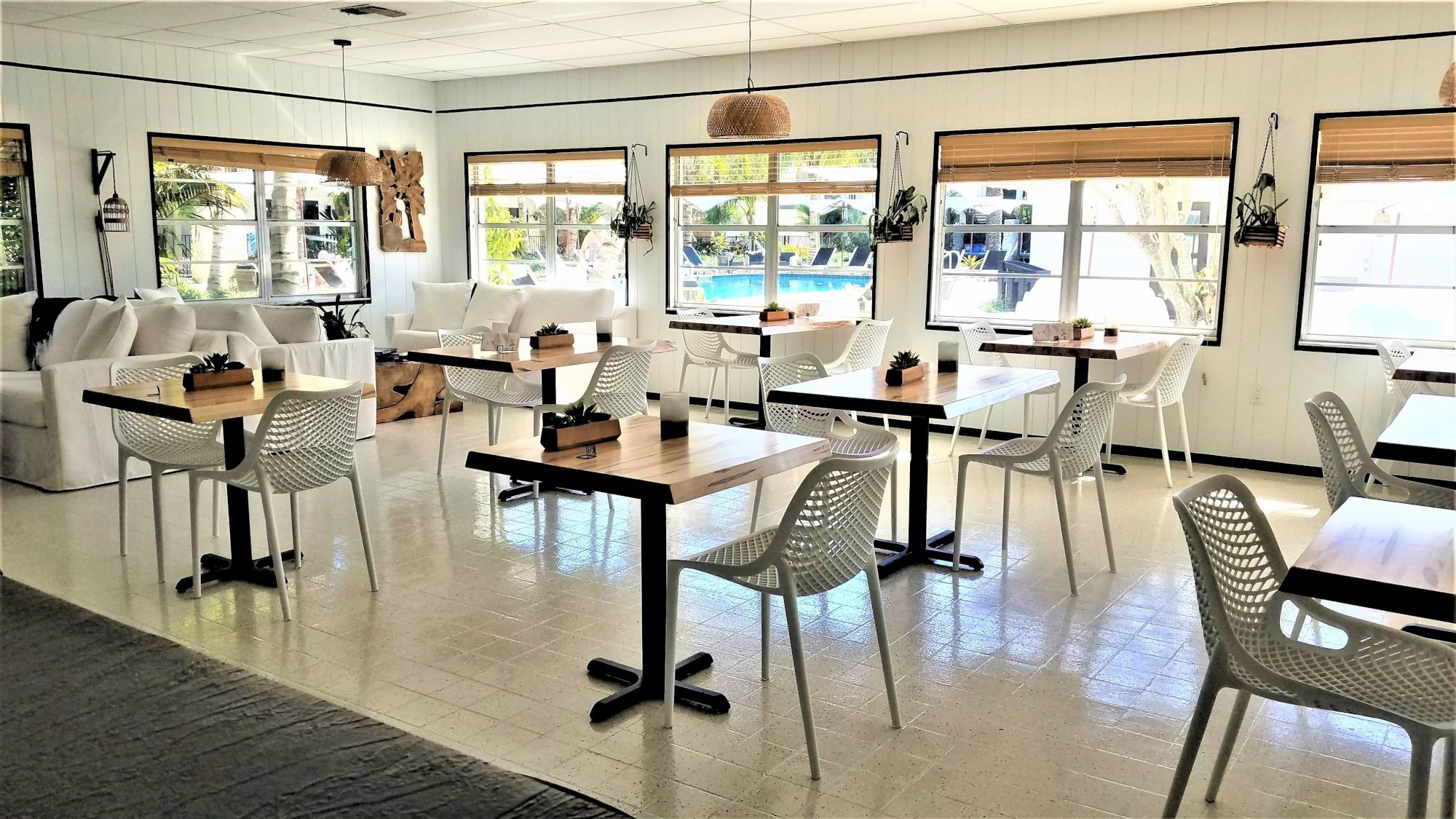 A bright, airy dining area with several tables and chairs, large windows, and minimalistic decor. Natural light floods the room, illuminating it.