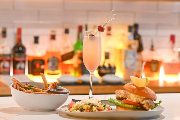 The image shows a bar counter with a drink, a bowl of fries and a burger with a side salad, set against a background of illuminated bottles.