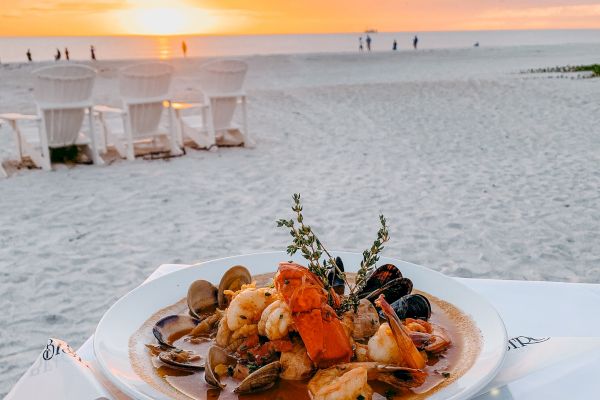 A plate of seafood pasta sits on a table by the beach at sunset, with the sea and sky in the background.