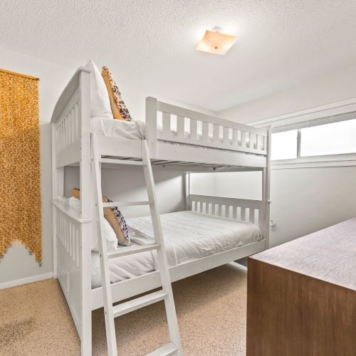 The image shows a small bedroom with white bunk beds, a wall hanging, a window, and a TV on a wooden dresser.