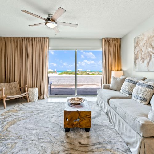 A cozy living room features a gray sofa, wooden coffee table, TV, light wood furniture, ceiling fan, large rug, and large window with ocean view.