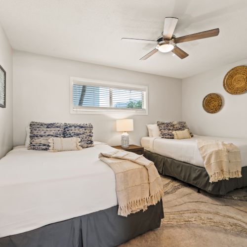 A bedroom with two beds, decorative pillows and blankets, a ceiling fan, wall art, a lamp, and a large mirror on the right wall.