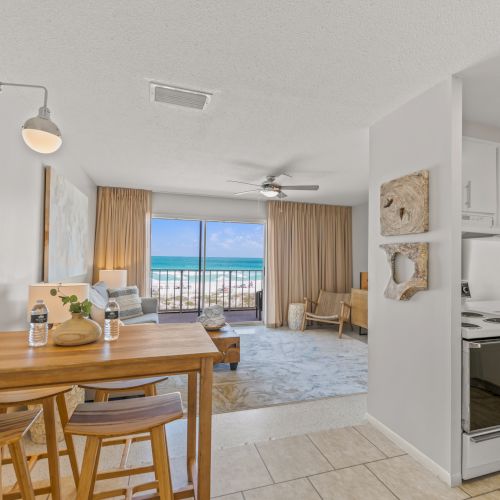 A bright coastal apartment features a dining area, kitchen, living room, and a large window with an ocean view, furnished simply and modernly ending the sentence.