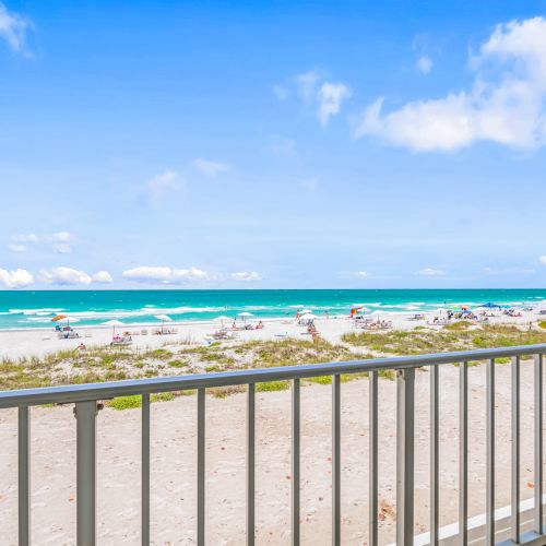 A view from a balcony overlooking a sandy beach with people, umbrellas, and clear blue skies above a turquoise ocean ending the sentence.