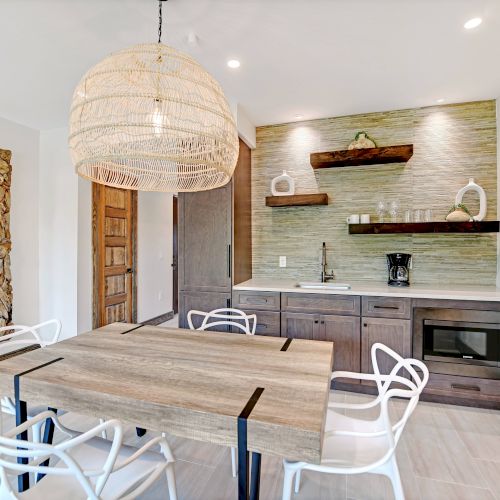 A modern dining area with a wooden table, white chairs, large wicker light fixture, shelves, and a kitchenette with a microwave and coffee maker.