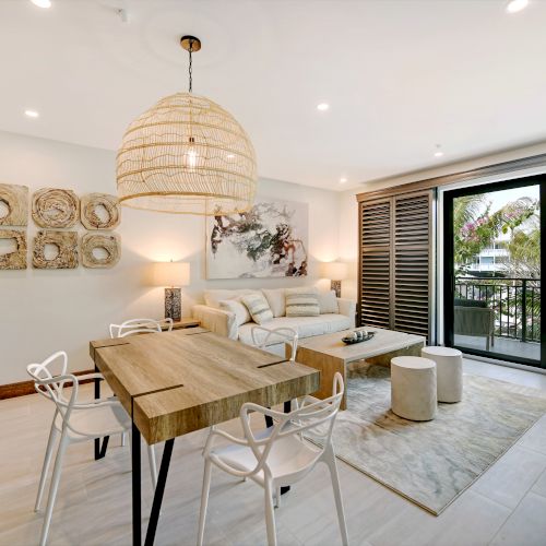 A modern living and dining area with a wooden table, white chairs, a rattan light fixture, a comfortable seating area, and large windows to a balcony and lush views.