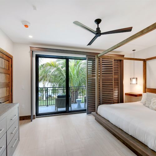 A chic bedroom with a canopy bed, dresser with TV, ceiling fan, and balcony overlooking tropical greenery through sliding glass doors.