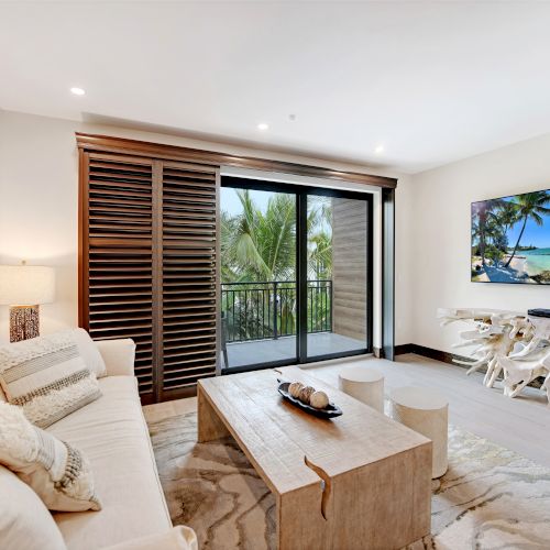 A modern living room with a sofa, coffee table, wall art, large TV, and sliding door opening to a balcony with tropical scenery outside ends the sentence.