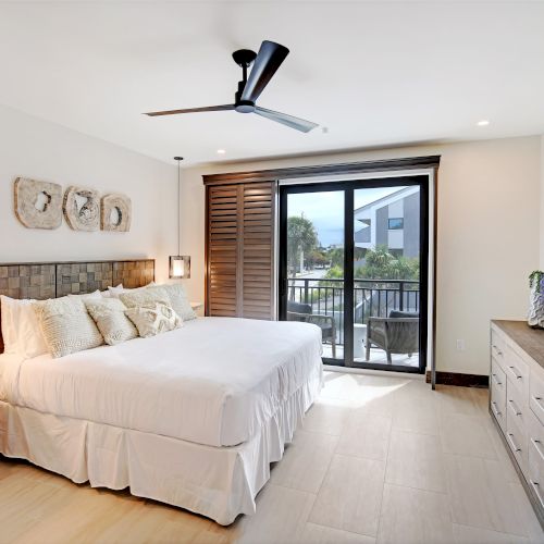 A modern bedroom features a large bed, stylish lamps, a ceiling fan, a flat-screen TV, and a sliding glass door with a balcony view, ending the sentence.