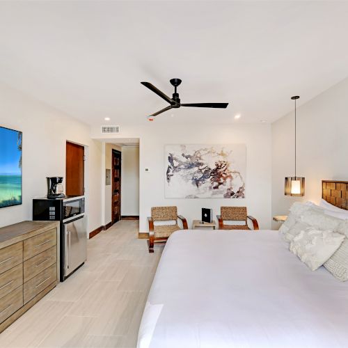 The image shows a modern bedroom with a bed, TV, mini fridge, and wall art. It has a ceiling fan, light-colored walls, and wood accents.