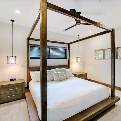 This image shows a modern bedroom with a wooden four-poster bed, two nightstands, hanging pendant lights, wall art, and a ceiling fan.