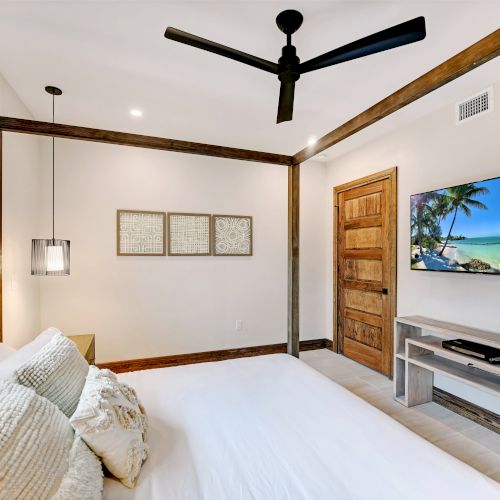 A cozy bedroom with a white bed, wooden accents, ceiling fan, wall TV displaying a beach scene, three framed art pieces, and a window.