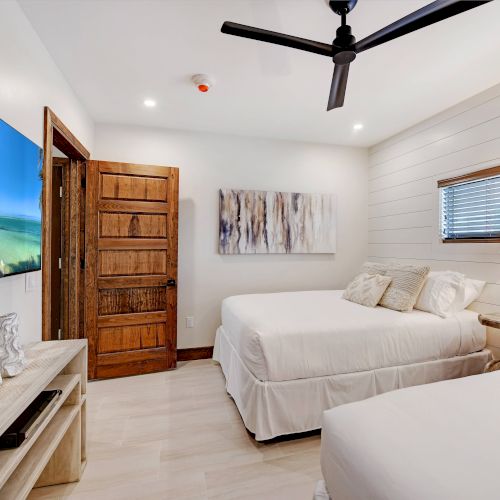 A cozy bedroom features two beds, a wooden door, a table lamp, artwork on the wall, a ceiling fan, and a TV showing a beach scene.