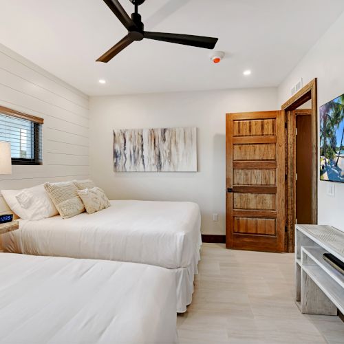 A cozy bedroom features two beds, a wooden door, mounted TV, a table, a tropical beach image on the TV, and a side table with decorations, ending the sentence.