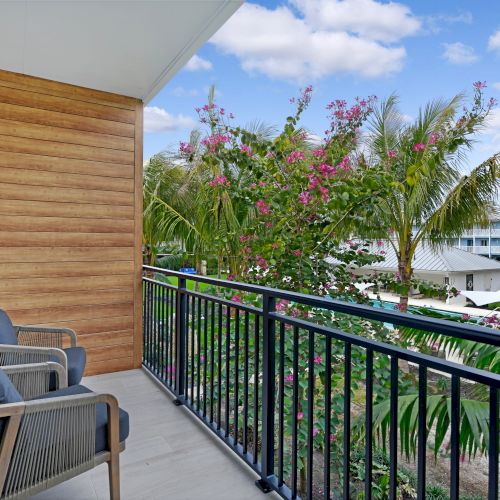 A balcony features two chairs with cushions overlooking a tropical garden with various trees and flowers, and a distant view of buildings.