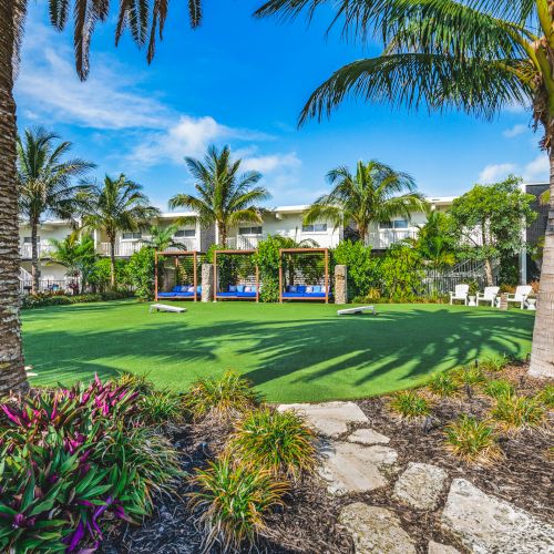 A lush, landscaped garden with palm trees, hammocks and lounge chairs, in front of a resort building under a clear blue sky.