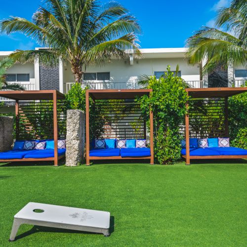 A lush outdoor area with three cabanas featuring blue cushions, palm trees, and a bean bag toss game on the grass.