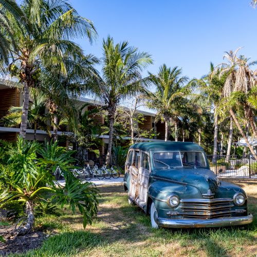 A retro car is parked on grass surrounded by tropical palm trees near a hotel or resort building under a clear blue sky.