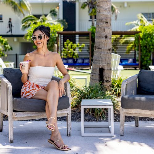 A person sits in an outdoor chair, holding a drink and wearing sunglasses. There are two empty chairs and palm trees in the background.