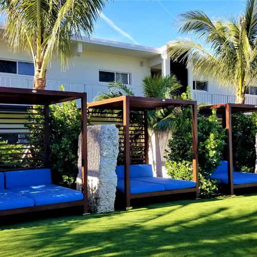 The image shows three outdoor cabanas with blue seating, surrounded by lush greenery and palm trees, in front of a modern building.